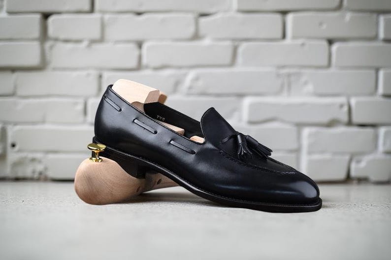 May include: Black leather loafer with a tassel detail, resting on a wooden shoe tree. The shoe has a sleek design and is set against a white brick wall background.