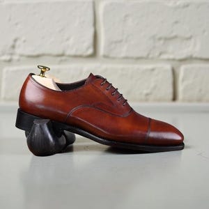 May include: A polished brown leather oxford shoe with a cap toe, dark brown laces, and a black sole. The shoe is supported by a wooden shoe tree with a gold-toned metal knob and rests on a dark, curved support piece, showcasing its profile.
