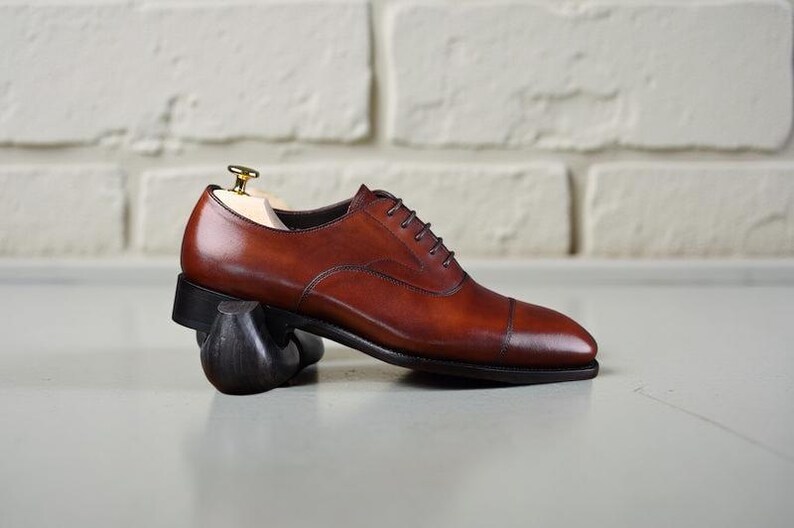May include: A polished brown leather oxford shoe with a cap toe, dark brown laces, and a black sole. The shoe is supported by a wooden shoe tree with a gold-toned metal knob and rests on a dark, curved support piece, showcasing its profile.