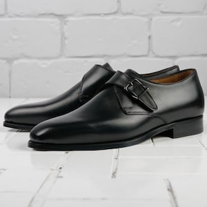 Könnte beinhalten: Ein Paar schwarze Lederschuhe mit einem Monkstrap-Schnallenverschluss. Die Schuhe haben ein schlankes, poliertes Aussehen und einen niedrigen Absatz. Die Schuhe werden auf einer weißen Oberfläche mit einem Backsteinmauer-Hintergrund präsentiert.