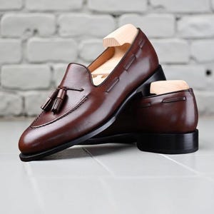 May include: A pair of brown leather loafers with tassel detailing. The shoes have a classic design with a rounded toe and a low heel. The shoes are displayed on a white tiled surface with a brick wall background.