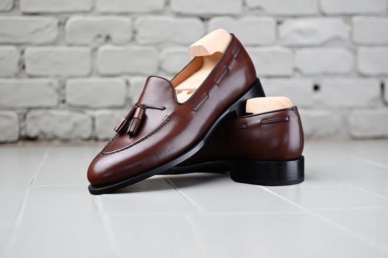 May include: A pair of brown leather loafers with tassel detailing. The shoes have a classic design with a rounded toe and a low heel. The shoes are displayed on a white tiled surface with a brick wall background.
