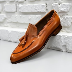 May include: A single tan leather loafer with a tassel. The shoe has a pointed toe and a stitched design around the edges. The heel is a dark brown colour. The shoe is a classic design.