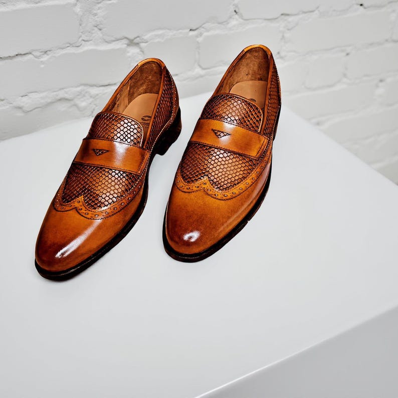 Pu&ograve; includere: Un paio di mocassini in pelle marrone con tomaia strutturata e design decorativo a punta di ala. Le scarpe hanno una finitura lucida e un cinturino in pelle sulla parte superiore. Le scarpe sono esposte su una superficie bianca.