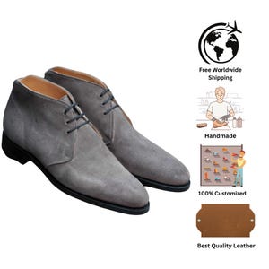 May include: A pair of gray suede chukka boots with black laces and soles. The boots have a classic design and are suitable for various occasions. The image also includes text that says "Handmade" and "Best Quality Leather".