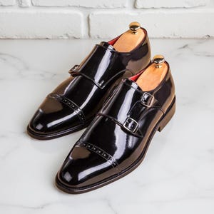 May include: A pair of black patent leather double monk strap shoes featuring a cap toe with decorative brogue detailing, silver-toned metal buckles, and a red interior lining, each shoe is fitted with a wooden shoe tree with a gold-toned knob.
