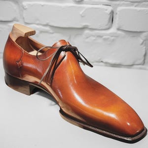 May include: A polished tan leather oxford shoe with a square toe, dark brown laces, and a wooden shoe tree inserted into the heel. The leather features a smooth, glossy finish with subtle color gradients and a small star-shaped detail on the side.