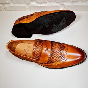 Pu&ograve; includere: Un paio di mocassini in pelle marrone con tomaia strutturata e finitura lucida. La suola di una scarpa &egrave; visibile, mostrando un battistrada in gomma nera. L'altra scarpa mostra la soletta con la scritta "Hand Made Only The Best Leather".
