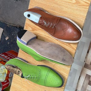 May include: A tan leather wingtip shoe with a white shoe tree, a shoe with a light green upper and a tan sole, and a bright lime green cap-toe shoe with white laces. These shoes feature decorative brogue perforations and lace-up closures.