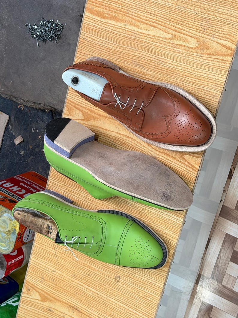 May include: A tan leather wingtip shoe with a white shoe tree, a shoe with a light green upper and a tan sole, and a bright lime green cap-toe shoe with white laces. These shoes feature decorative brogue perforations and lace-up closures.