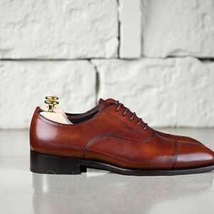 May include: A polished brown leather oxford shoe with a cap toe design, dark brown laces, and a low stacked heel. A light-coloured wooden shoe tree with a gold-toned metal knob is inserted into the opening of the shoe to maintain its shape.
