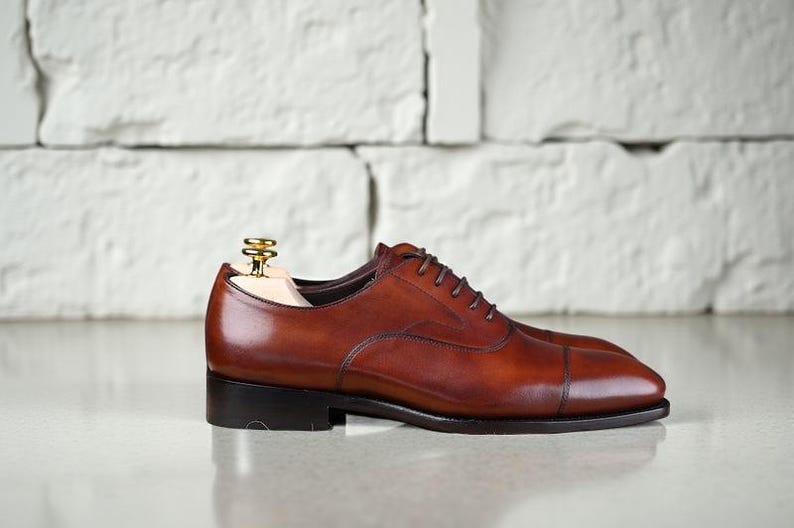 May include: A polished brown leather oxford shoe with a cap toe design, dark brown laces, and a low stacked heel. A light-coloured wooden shoe tree with a gold-toned metal knob is inserted into the opening of the shoe to maintain its shape.