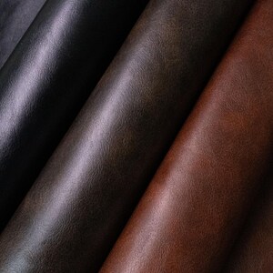 May include: Three rolls of textured material arranged diagonally. The leftmost roll is black, the center roll is a dark charcoal gray, and the rightmost roll is a rich reddish-brown. Each roll displays a consistent, fine-grained surface texture.