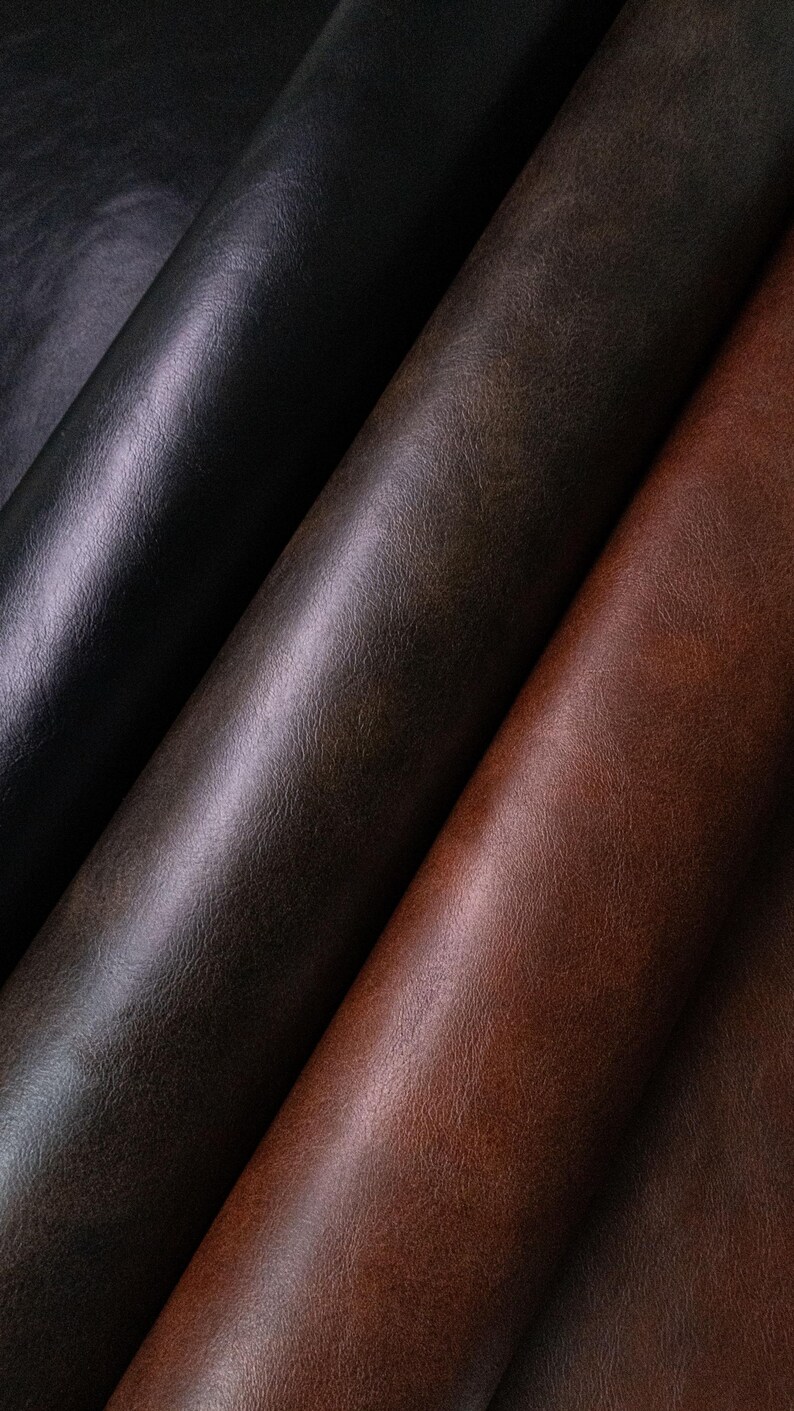 May include: Three rolls of textured material arranged diagonally. The leftmost roll is black, the center roll is a dark charcoal gray, and the rightmost roll is a rich reddish-brown. Each roll displays a consistent, fine-grained surface texture.