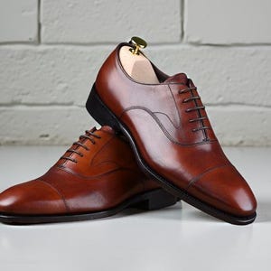 May include: A pair of brown leather oxford shoes with a cap toe design, dark brown laces, and black soles. One shoe features a light-coloured wooden shoe tree inserted inside, and both shoes have a polished finish with visible stitching details.