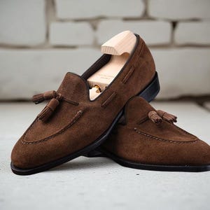 May include: A pair of dark brown suede loafers featuring decorative tassels on the vamp and a stitched apron toe. The shoes have a dark sole and are equipped with a light-colored wooden shoe tree inserted into the primary loafer for shape.