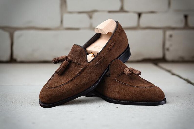 May include: A pair of dark brown suede loafers featuring decorative tassels on the vamp and a stitched apron toe. The shoes have a dark sole and are equipped with a light-colored wooden shoe tree inserted into the primary loafer for shape.