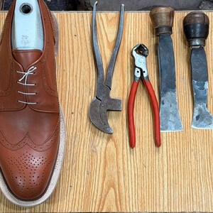 May include: A tan leather wingtip oxford shoe with white laces and a light-colored shoe tree insert, positioned next to a collection of metal shoemaking tools including pliers, a nipper, two flat-bladed knives, a metal awl, and a hammer.