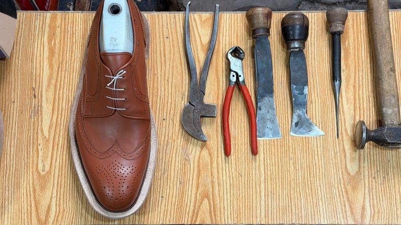 May include: A tan leather wingtip oxford shoe with white laces and a light-colored shoe tree insert, positioned next to a collection of metal shoemaking tools including pliers, a nipper, two flat-bladed knives, a metal awl, and a hammer.