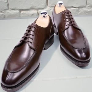 Op de afbeelding: Een paar bruine leren herenschoenen met een klassiek ontwerp. De schoenen hebben een vetersluiting, een ronde neus en een gepolijste afwerking. De schoenen worden getoond met houten schoenspanners erin.