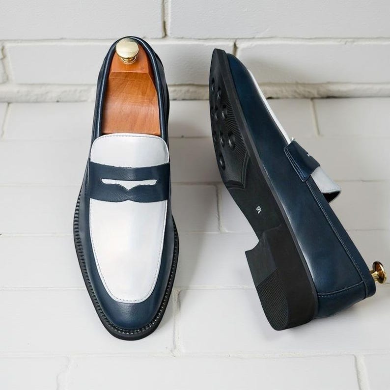 May include: Navy blue and white leather loafers. The shoes have a white vamp and a navy blue saddle, with a black sole. The shoes are displayed with a wooden shoe tree. The loafers are approximately 28 cm long.