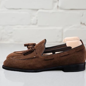 May include: A pair of dark brown suede loafers featuring decorative tassels on the vamp, a low black heel, and a rounded toe. The shoes are equipped with light-colored wooden shoe trees inserted inside to maintain their shape and structure.