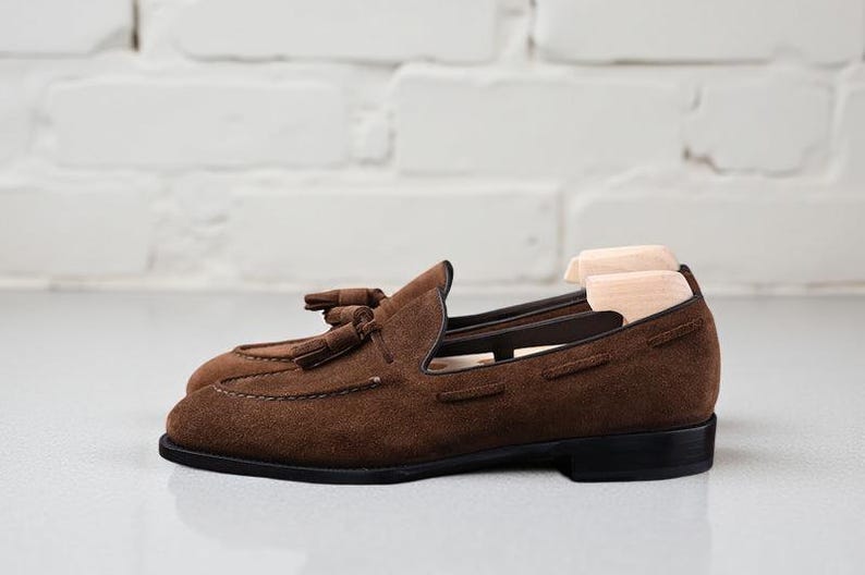 May include: A pair of dark brown suede loafers featuring decorative tassels on the vamp, a low black heel, and a rounded toe. The shoes are equipped with light-colored wooden shoe trees inserted inside to maintain their shape and structure.
