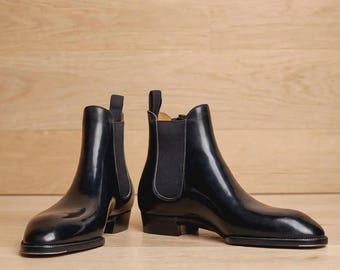 May include: A pair of black leather Chelsea boots with a polished finish, featuring elastic side panels, a rounded toe, a low stacked heel, and a fabric pull tab at the top of the ankle collar. The boots have a sleek silhouette and a smooth construction.