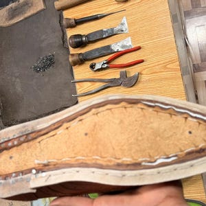 May include: A leather shoe sole in the process of being crafted, featuring visible stitching along the perimeter and a textured tan center. Nearby tools include metal pliers, chisels, and a wooden handle, all arranged on a wooden workbench.