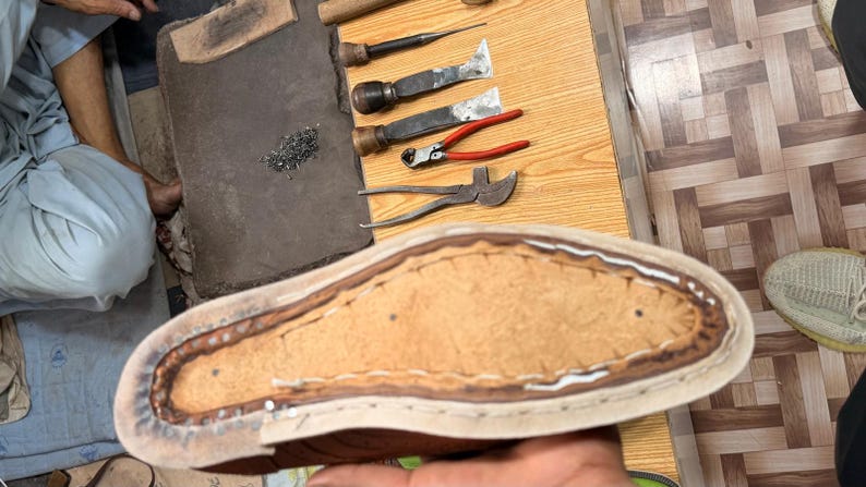 May include: A leather shoe sole in the process of being crafted, featuring visible stitching along the perimeter and a textured tan center. Nearby tools include metal pliers, chisels, and a wooden handle, all arranged on a wooden workbench.