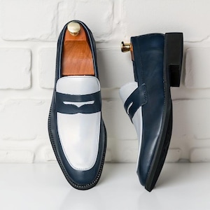 May include: Two-tone leather loafers in navy blue and white. The shoes have a navy blue upper, a white vamp, and a navy blue strap. The soles are black. The shoes are displayed with wooden shoe trees.