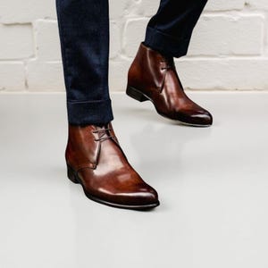 May include: Brown leather ankle boots with a lace-up design. The boots have a polished finish and are worn with dark blue trousers. The boots are a classic style, suitable for various occasions.