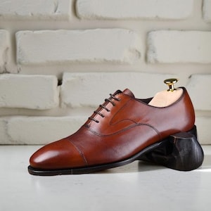May include: A polished cognac brown leather oxford shoe with a cap toe design, featuring dark brown laces and a slim dark sole. A light-coloured wooden shoe tree with a gold-toned metal knob is inserted into the heel area of the footwear.