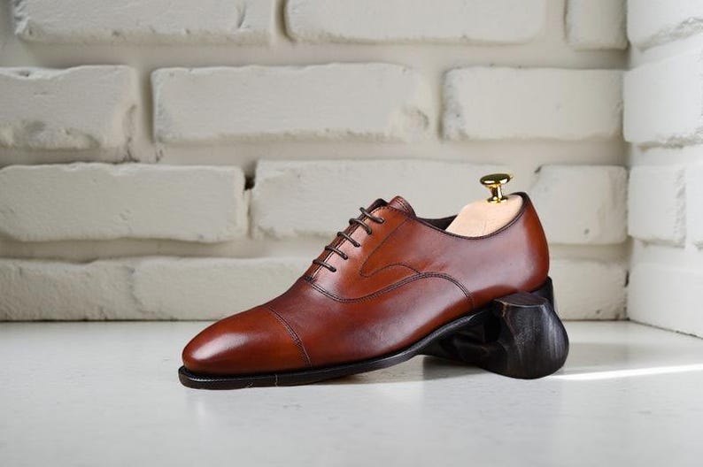 May include: A polished cognac brown leather oxford shoe with a cap toe design, featuring dark brown laces and a slim dark sole. A light-coloured wooden shoe tree with a gold-toned metal knob is inserted into the heel area of the footwear.