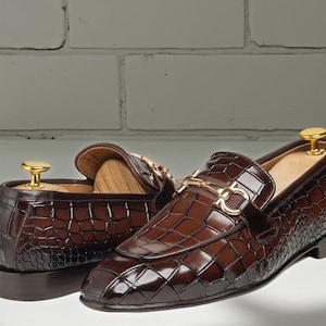 Puede incluir: Un par de mocasines de cuero marrón con un patrón de piel de cocodrilo. Los zapatos tienen una hebilla dorada y una horma de madera en el interior. Los mocasines se exhiben sobre una superficie blanca.