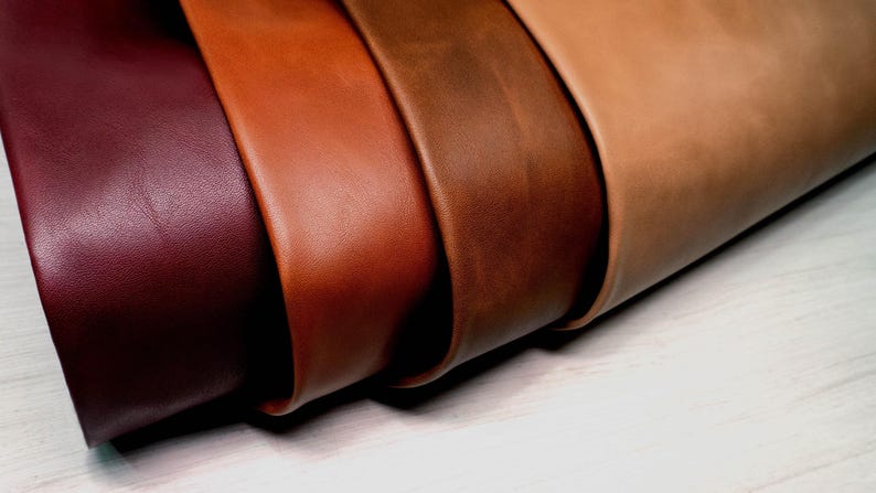 May include: Four distinct sheets of smooth leather arranged in a gradient of colors, starting with a deep burgundy on the left, followed by a vibrant orange-brown, a rich chocolate brown, and ending with a light tan shade on the far right side.