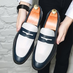 May include: Two-tone leather loafers in navy blue and white. The shoes have a navy blue saddle strap and heel, with a white vamp. The soles are black with a stitched border. The shoes are held by a person.