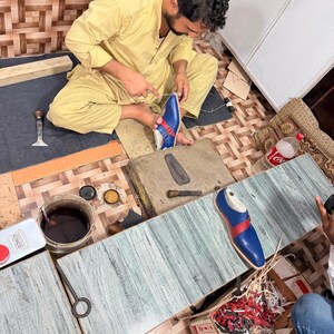 May include: A blue leather shoe with a red strap and silver buckle being hand-stitched by a person. The shoe features a pointed toe and a light-colored sole. Nearby are various shoemaking tools, including a metal tool, a container of dark liquid, and a box filled with leather scraps.