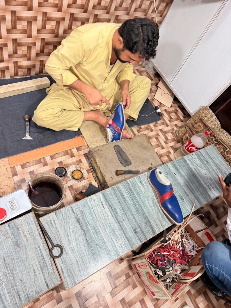 May include: A blue leather shoe with a red strap and silver buckle being hand-stitched by a person. The shoe features a pointed toe and a light-colored sole. Nearby are various shoemaking tools, including a metal tool, a container of dark liquid, and a box filled with leather scraps.