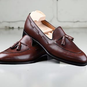May include: A pair of brown leather loafers with tassel detailing. The shoes have a classic design with a rounded toe and a low heel. Shoe trees are inserted inside the shoes. The loafers are displayed on a white surface.