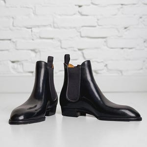 May include: A pair of black leather Chelsea boots with elastic side panels. The boots have a sleek, polished appearance and a low heel. The background is a white brick wall and a white floor.