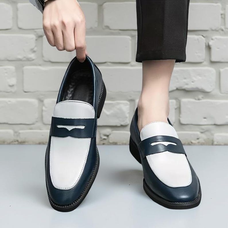 May include: Navy blue and white leather loafers. These shoes feature a classic penny loafer design with a rounded toe and a low heel. The upper is navy blue with a white vamp and a decorative strap. The shoes are designed for comfort and style, suitable for various occasions.