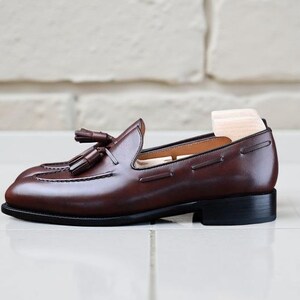 May include: A brown leather loafer with tassel detailing. The shoe features a low heel and a wooden shoe tree. The loafer is displayed on a white tiled surface.