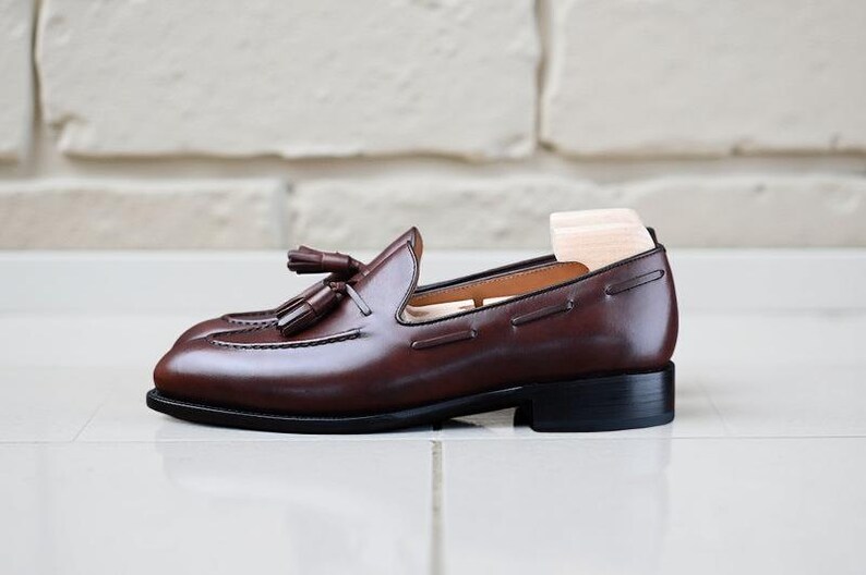 May include: A brown leather loafer with tassel detailing. The shoe features a low heel and a wooden shoe tree. The loafer is displayed on a white tiled surface.
