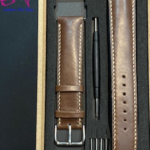 May include: A brown leather watch strap with white stitching and a silver buckle is displayed in a box with a tool for changing watch straps.