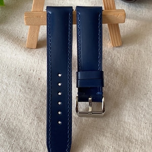 Puede incluir: Dos correas de reloj de cuero azul marino con costuras blancas. Una correa tiene una hebilla plateada. Las correas se muestran sobre una superficie de color claro. Las correas son de Leather Art Shop.