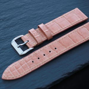 Puede incluir: Una correa de reloj de color rosa claro con un patrón texturizado de estilo caimán y costuras en color beige. La correa cuenta con una hebilla de metal plateado pulido y dos trabillas de cuero a juego. Incluye una serie de orificios perforados.