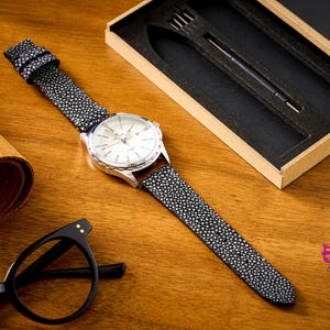 Könnte beinhalten: Eine silberne Armbanduhr mit weißem Zifferblatt und einem schwarz-weiß gesprenkelten Armband liegt auf einer Holzoberfläche. Ein aufgerolltes Stück braunes Leder und eine schwarze Brille sind ebenfalls zu sehen. Eine schwarze Box mit Stift und Gabel befindet sich im Hintergrund.