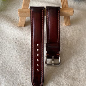 Peut inclure: Deux bracelets de montre en cuir marron foncé avec une finition polie. Les bracelets ont des coutures contrastées sur les bords et une boucle argentée. Les bracelets sont présentés sur une surface de couleur neutre.