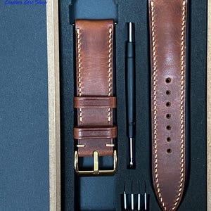 May include: A brown leather watch strap set with a gold-colored buckle, cream stitching, and a matching strap. The set includes a tool and pins, all presented in a black box. The text "Leather Art Shop" is visible in the upper left corner.